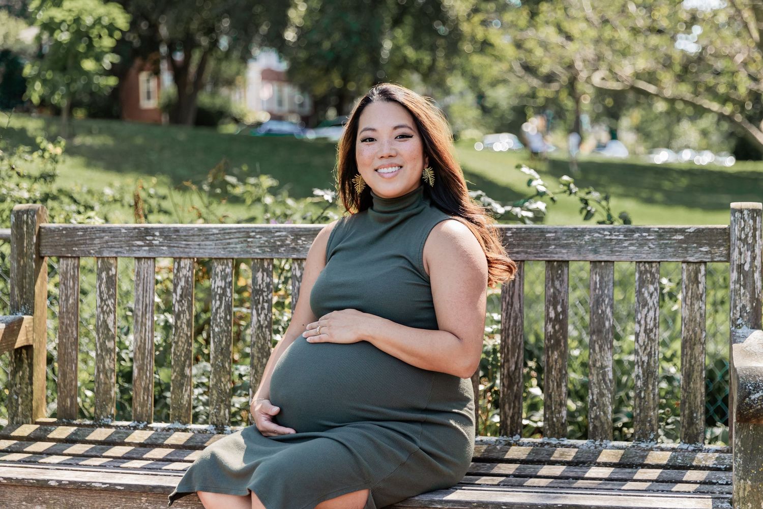 Young asian woman sits on a wooden bench as she wraps her hands around her baby bumps as the summer sun glistens through her hair