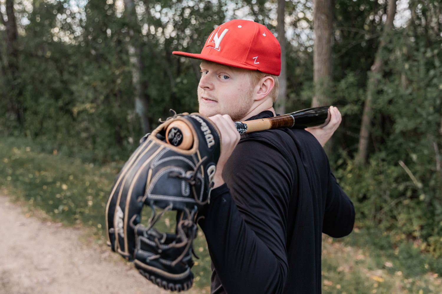 Cade rests his baseball bat across his shoulders with his glove hanging off the end of the bat as he stares into the camera