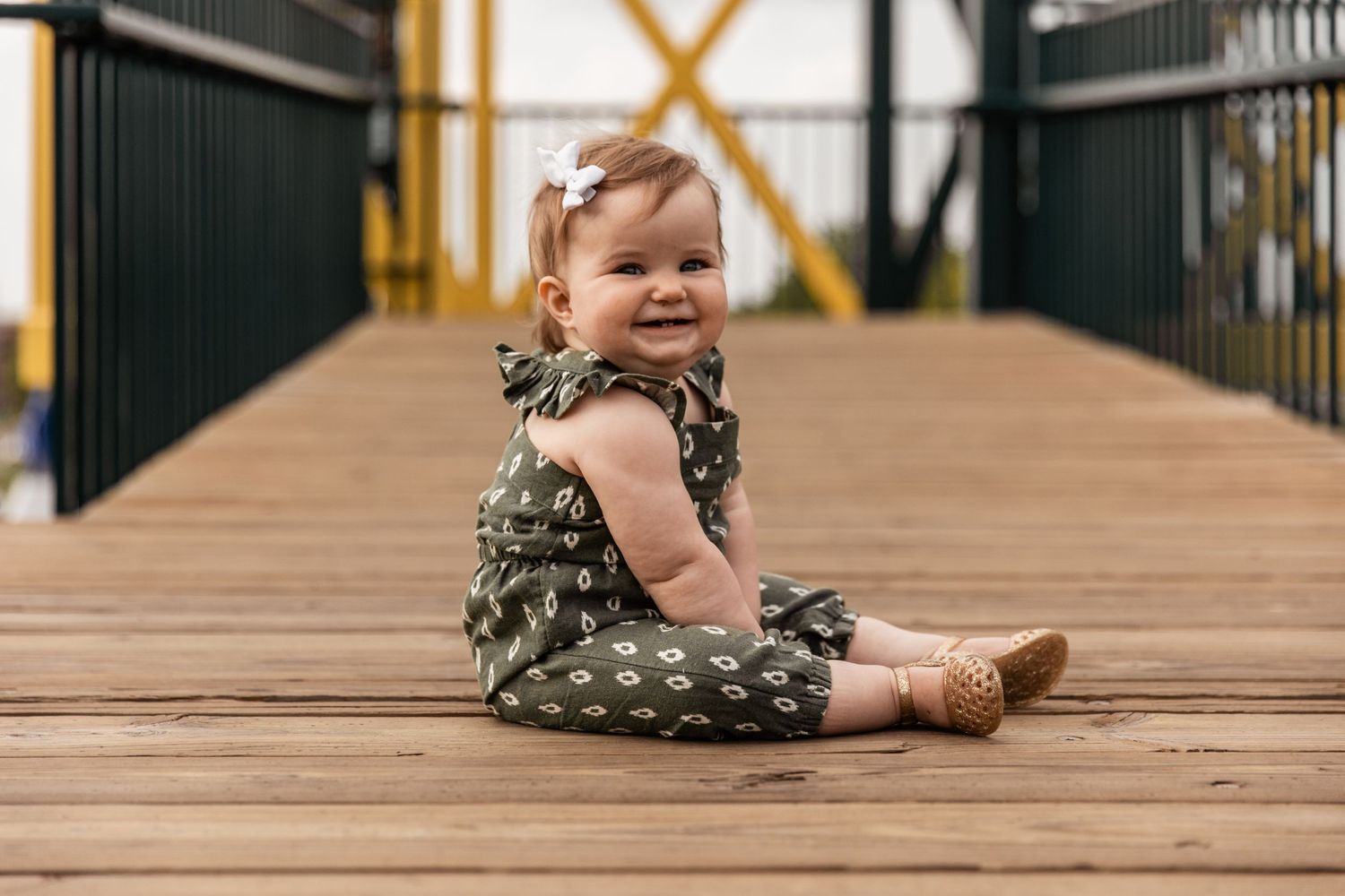 Adorable baby girl smiles and sits on a wooden and metal walkway leading up towards a bridge