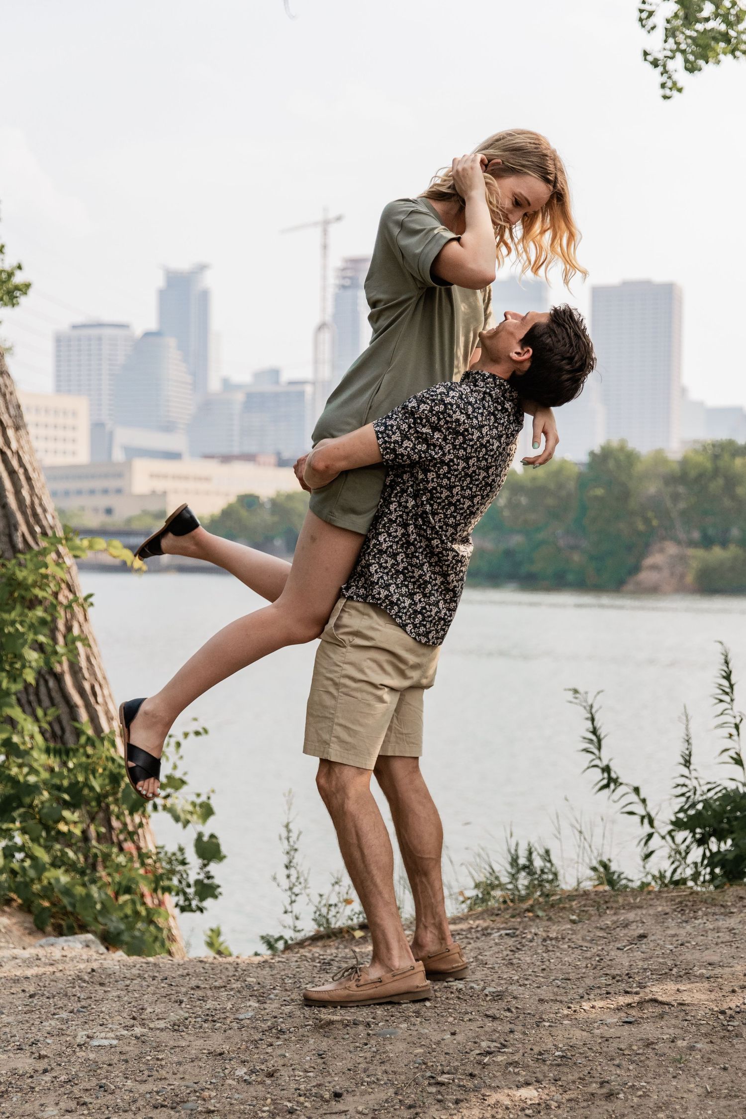young man lifts his girlfriend up with the minneapolis skyline behind them
