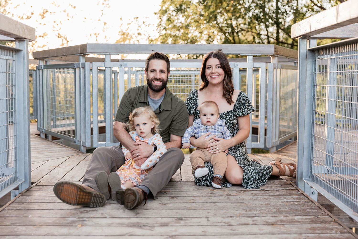 Family with two young children lean in towards each other with the kids on their laps as they sit on an overlook bridge