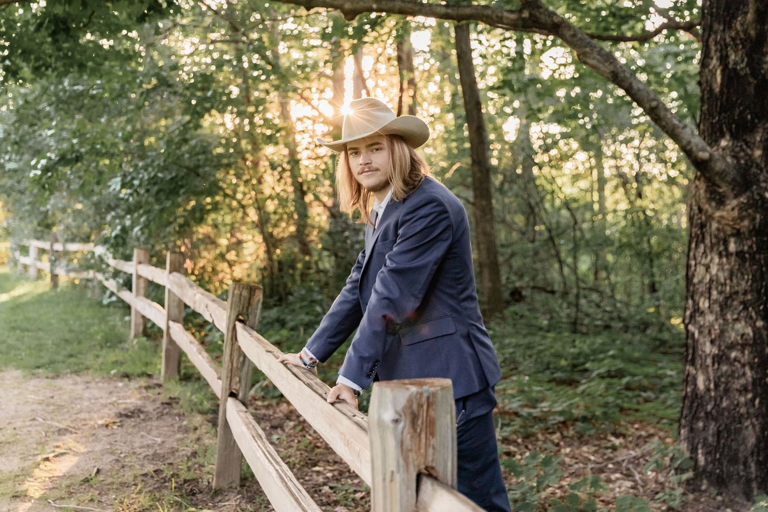 Young man leans over a wooden fence as the sun glows behind his cowboy hat at Carver Lake Park in Woodbury
