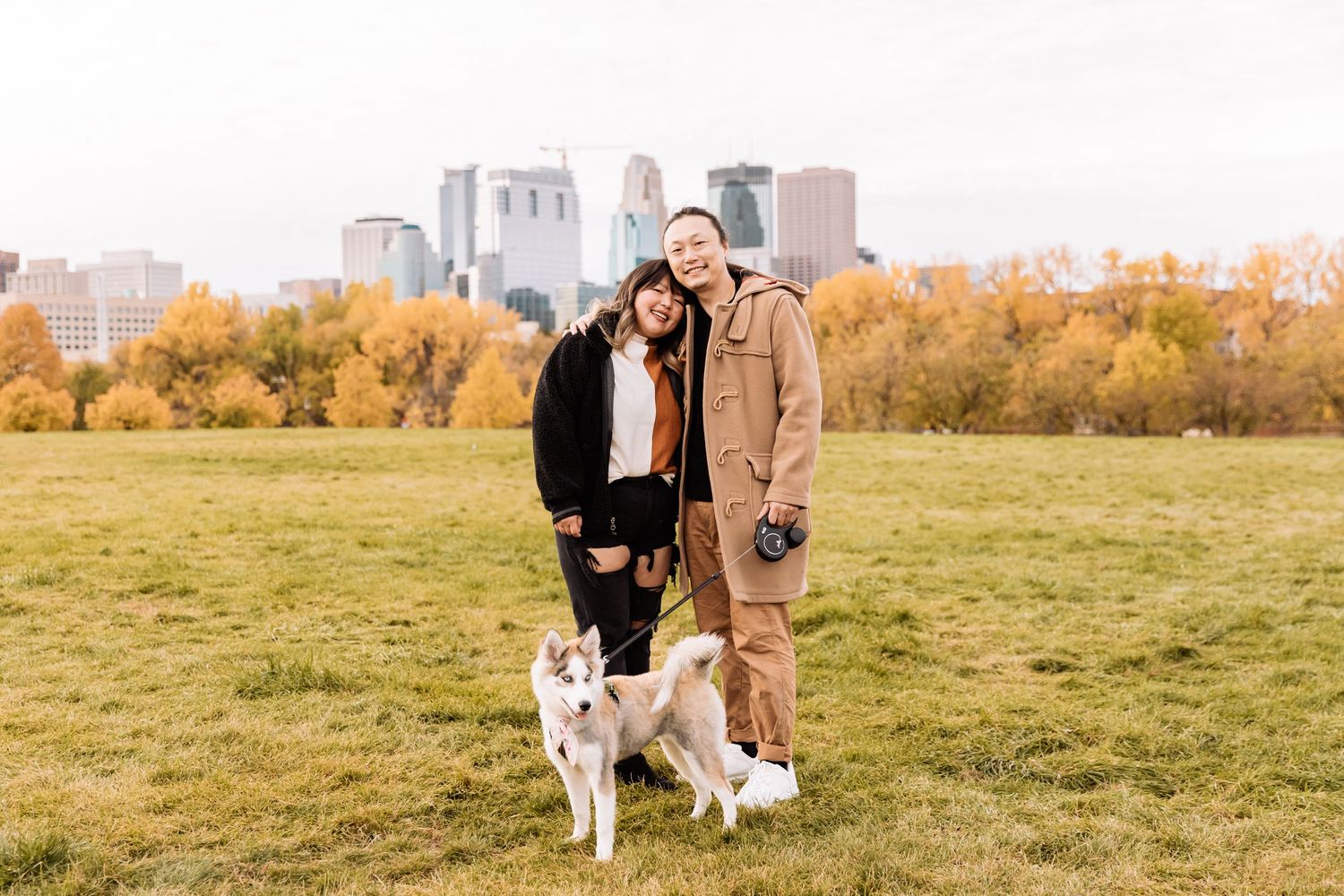 Asian couple stands with their husky in a grassy field at Boom Island Park