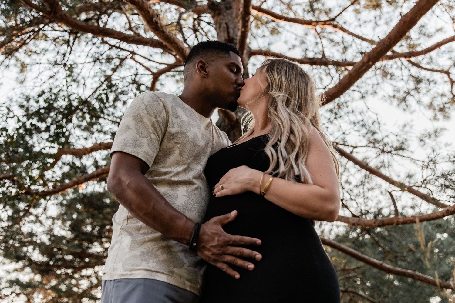 couple kisses under a large pine tree to announce their pregnancy at Boom Island Park
