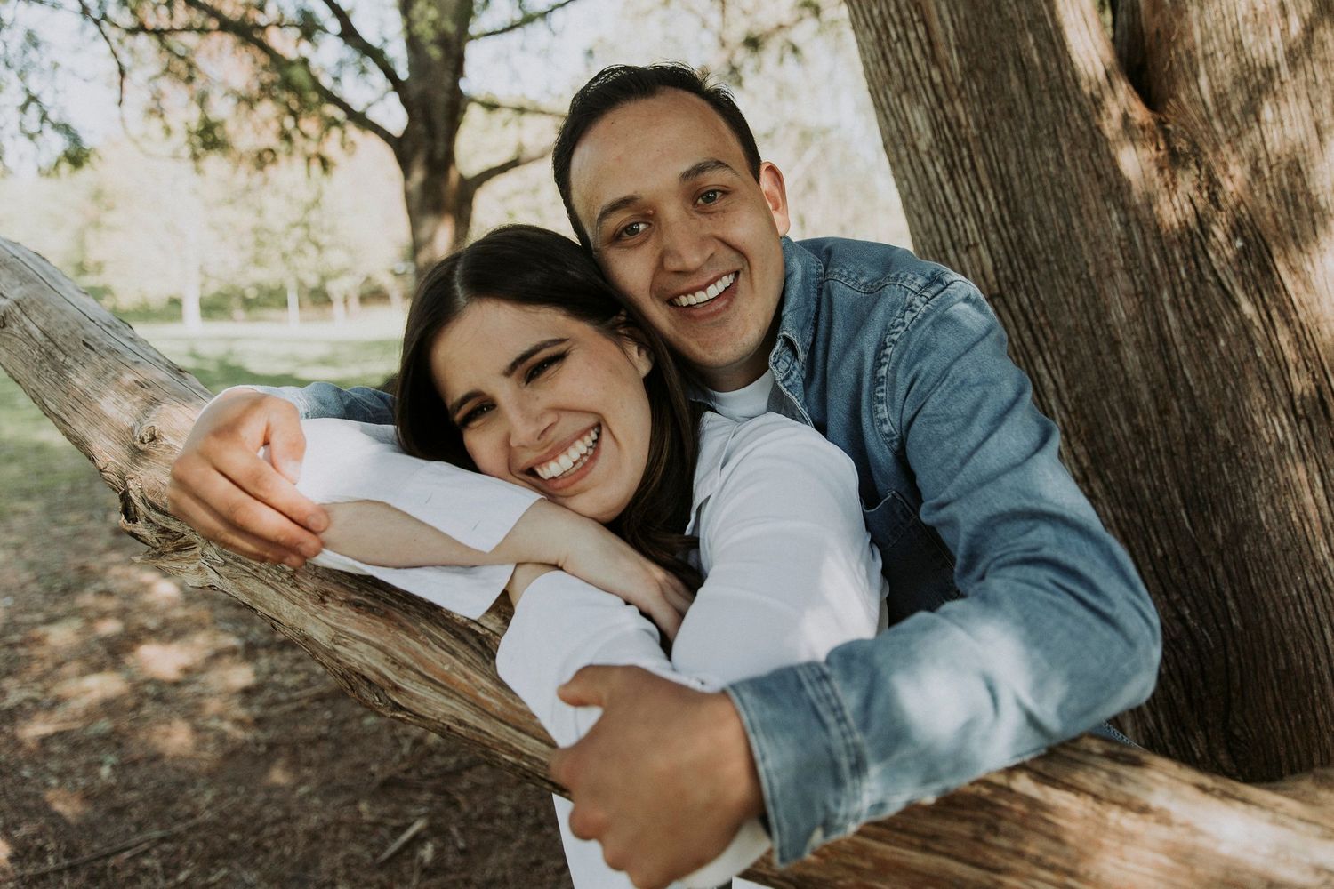 Young man wraps his arms around his fiance as they lean over a large tree branch