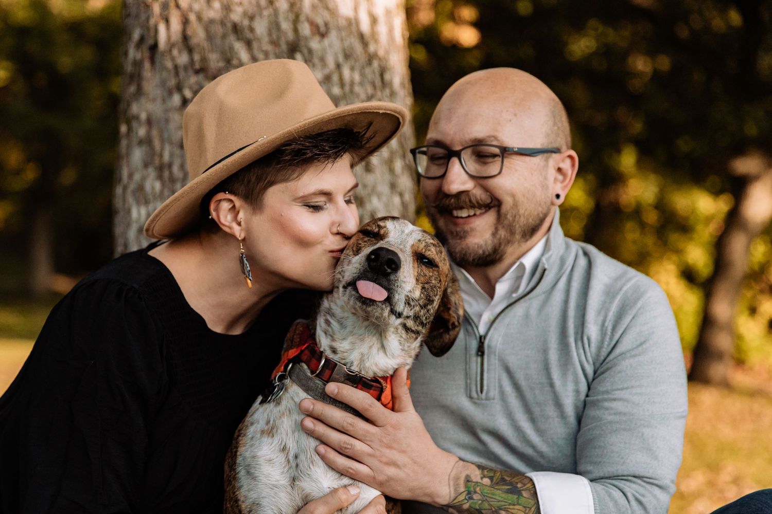 Husband and wife sit next to an oak tree with their puppy and kiss it on the head as it sticks it's tongue out