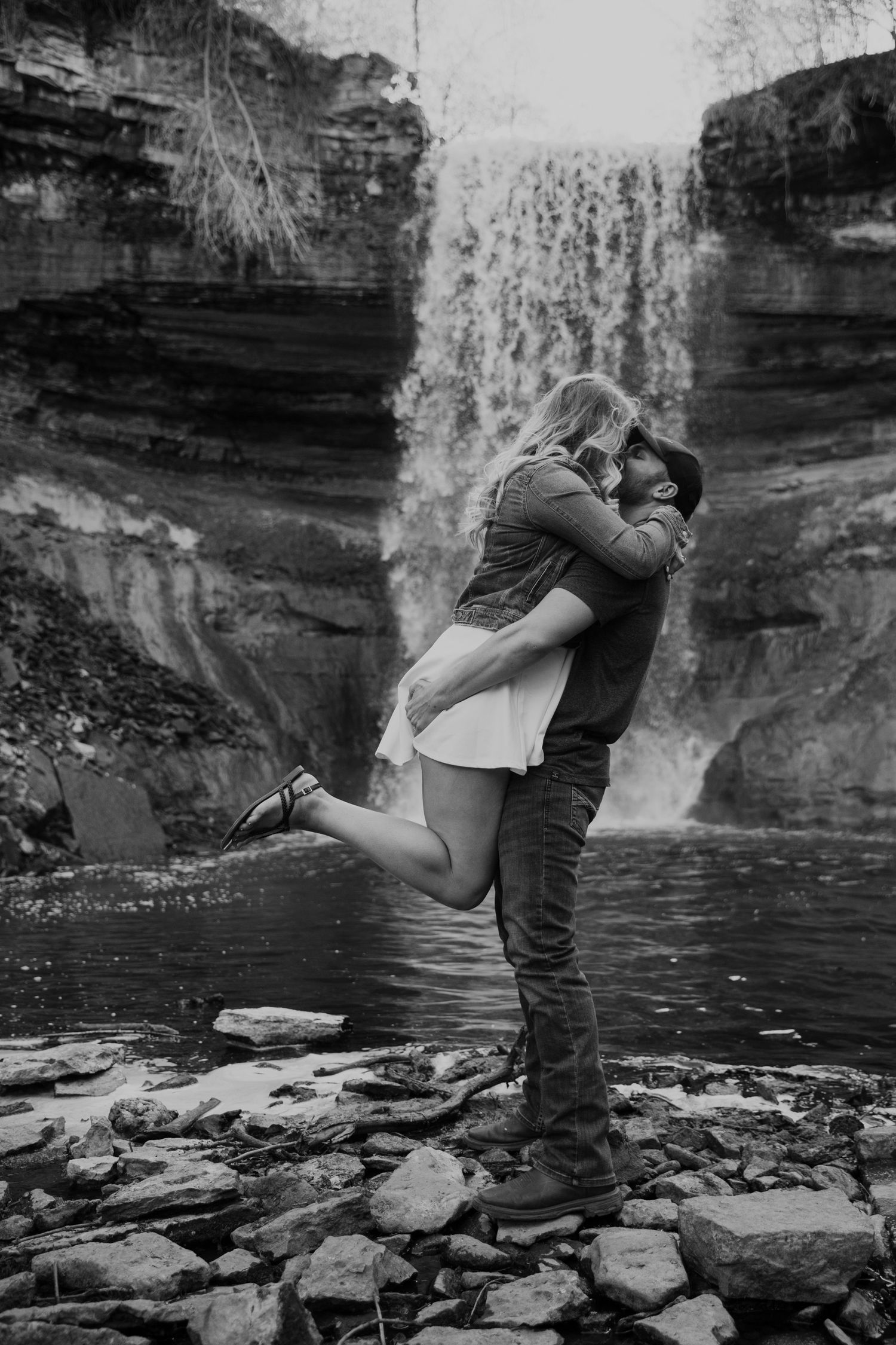young man lifts his fiance up for a kiss in front of a waterfall