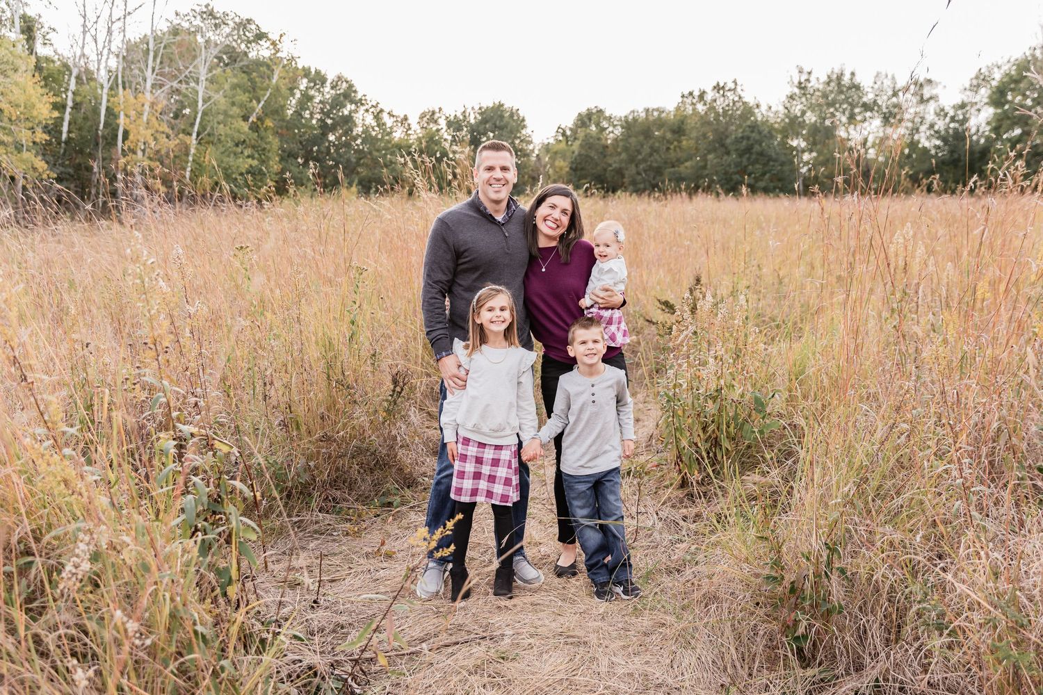 Greener family stands in a tall grassy field with fall colored trees surrounding them