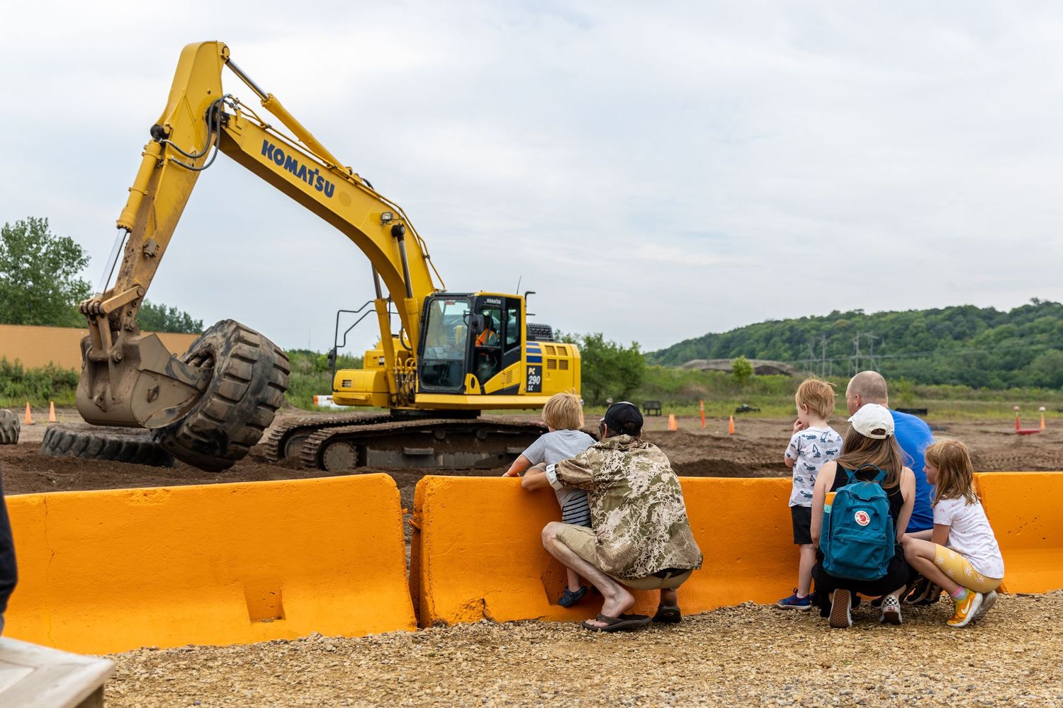 Construction equipment scoops a bucket full of dirt as kids and parents look on