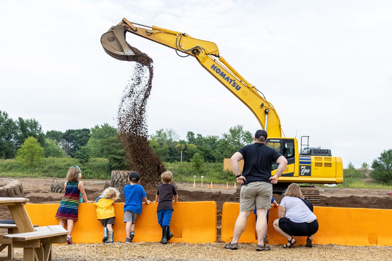 Back loader dumps an entire buck of dirt from up high as little kids watch