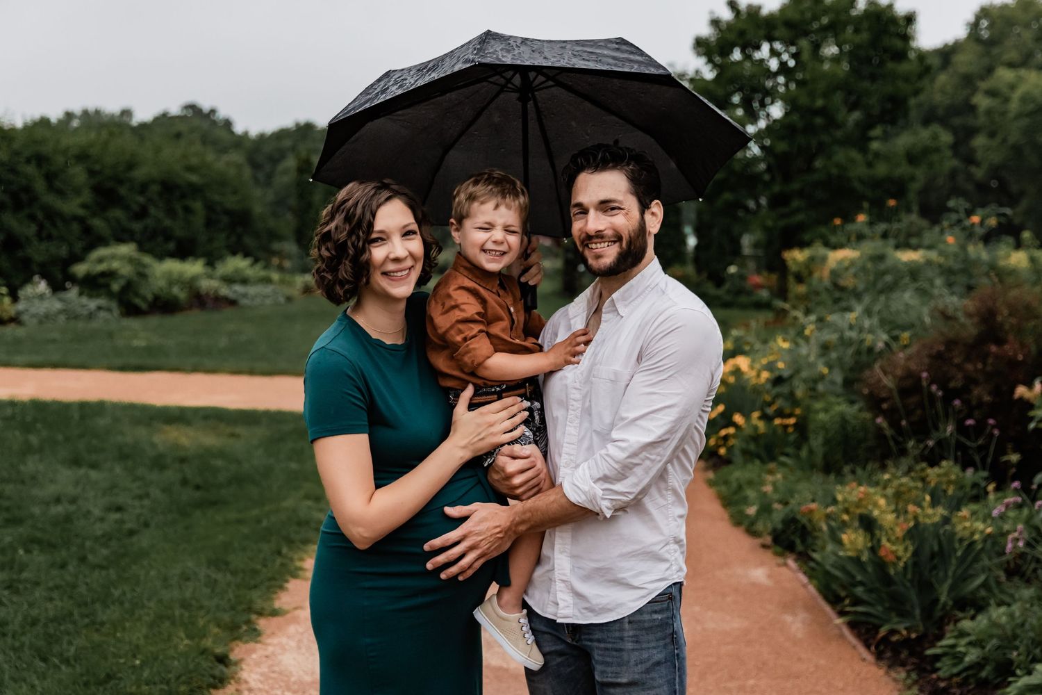 Pregnant wife and husband hold their son and an umbrella as the rain comes down on them