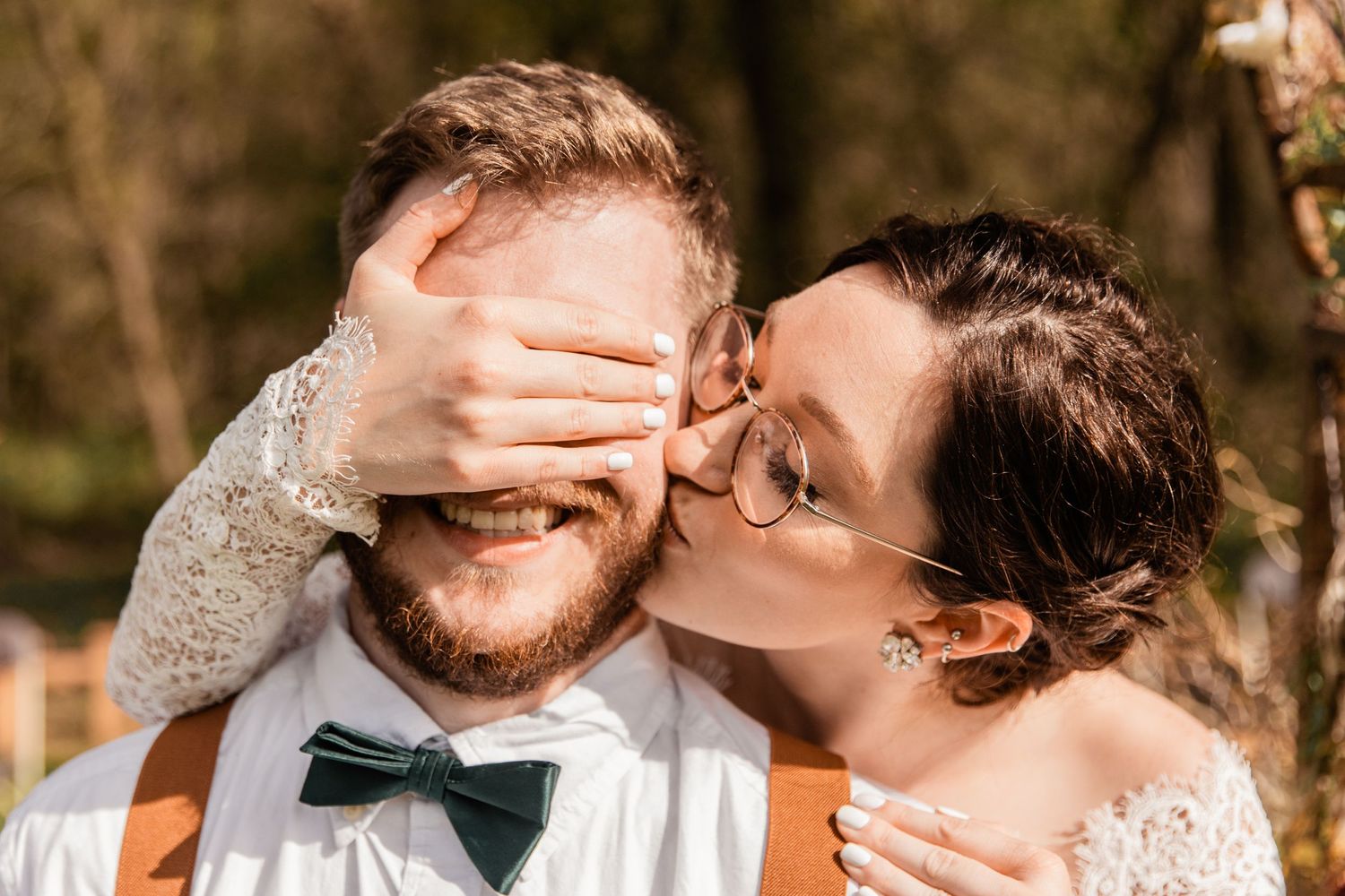 bride covers her groom's eyes and kisses him on the cheek before his first look