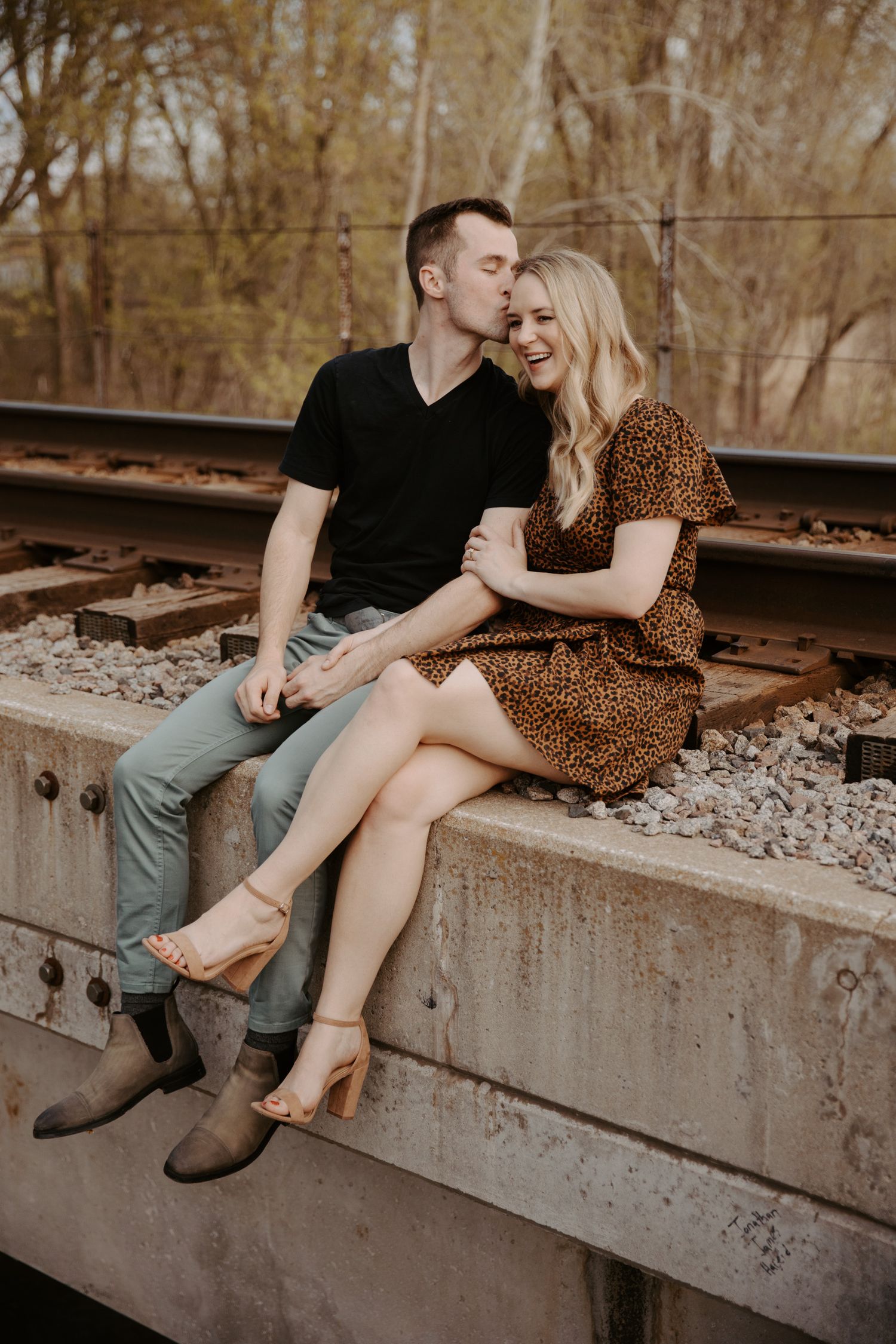 Young man kisses his fiance on her forehead as they sit on a bridge with their feet dangling over the side