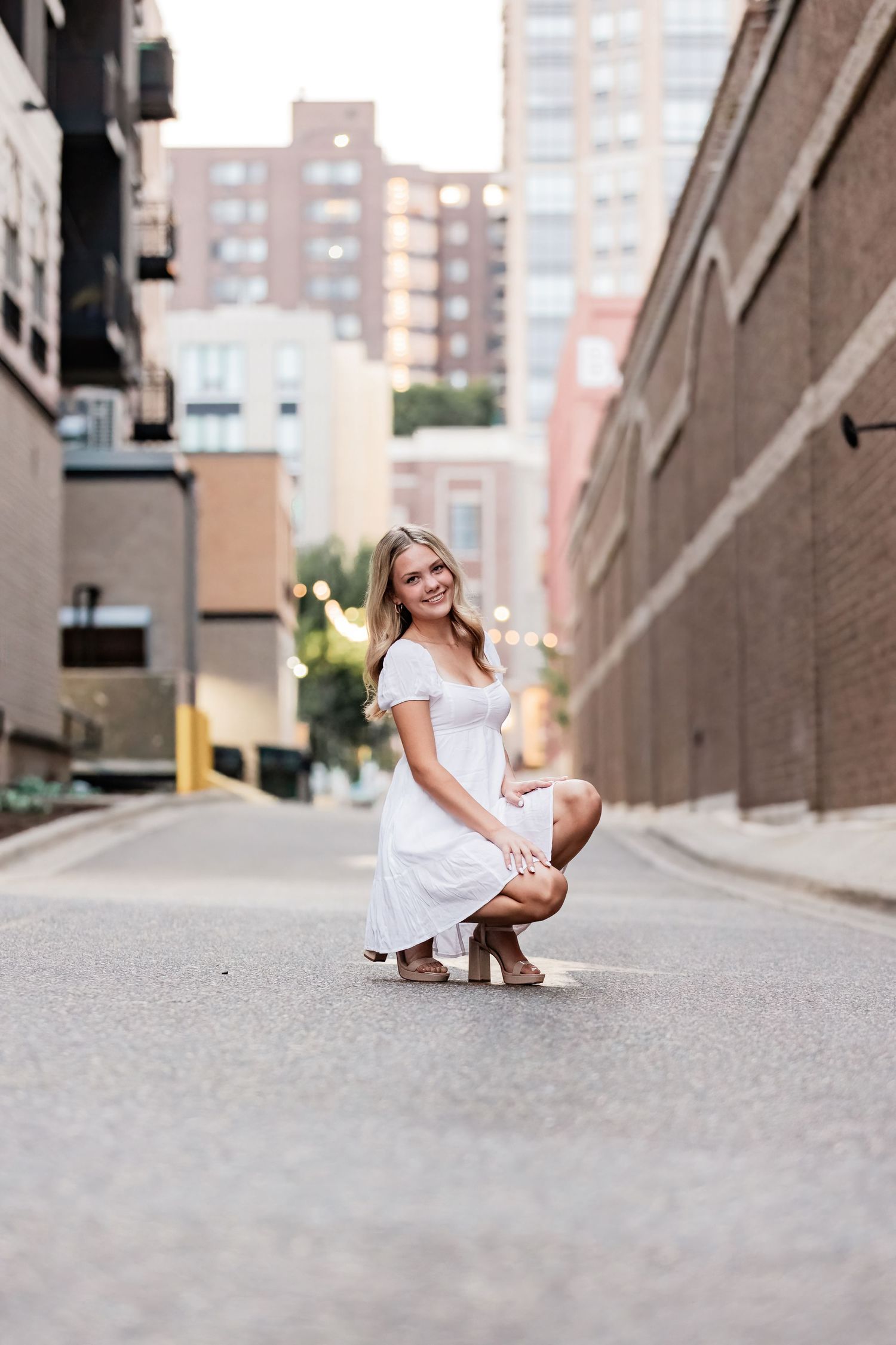 Lucy crouches down resting her hands on her thighs in a brick laden alley way