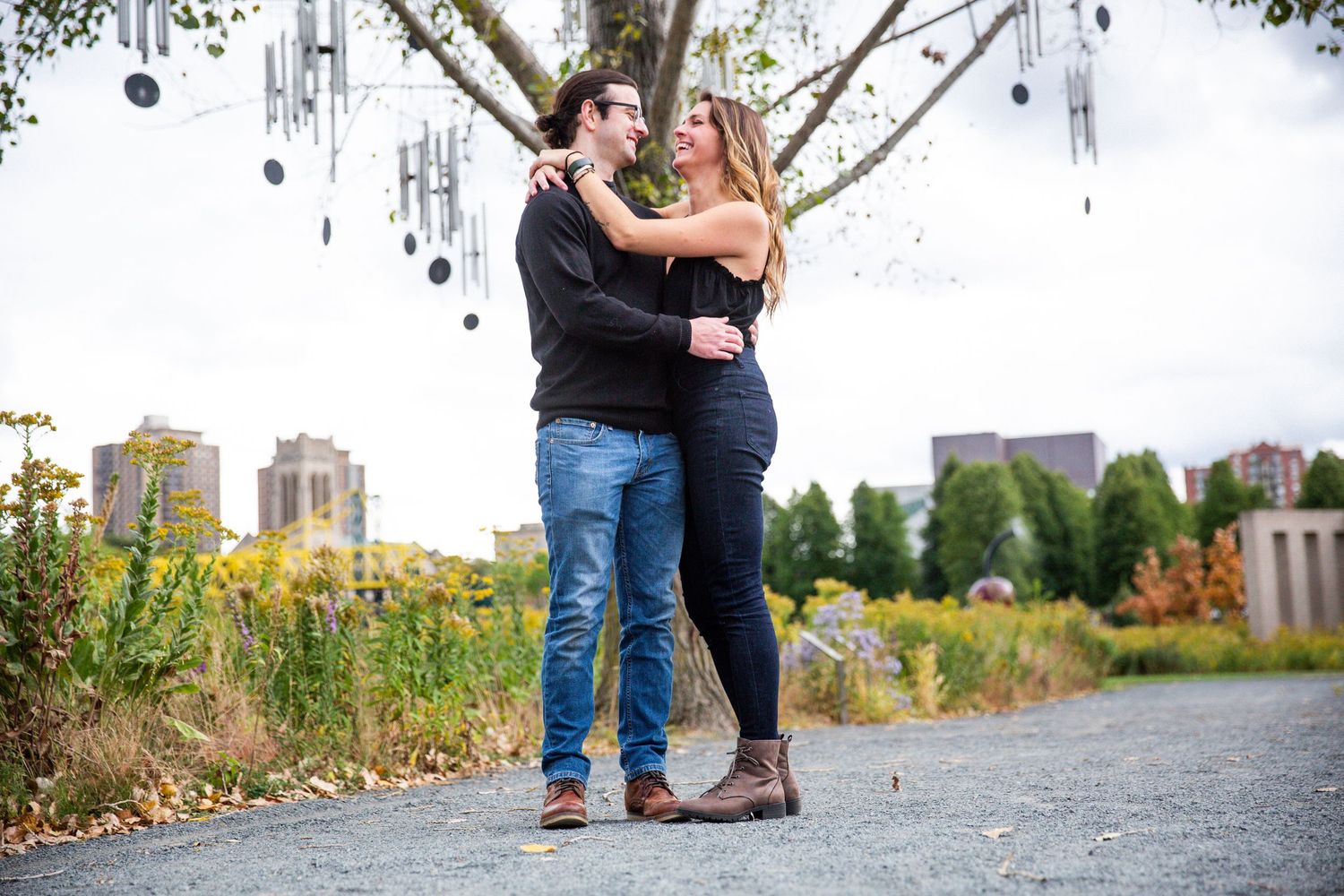 Young couple wraps their arms around each other under a tree full of wind chimes on a warm sunny day