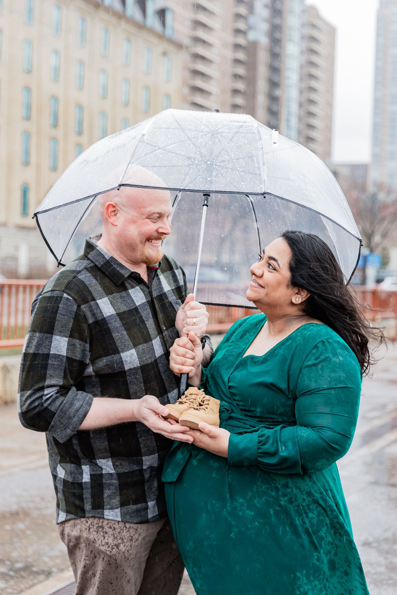 Husband and wife cozy up under a clear umbrella as the rain falls on them while they celebrate announcing their pregnancy