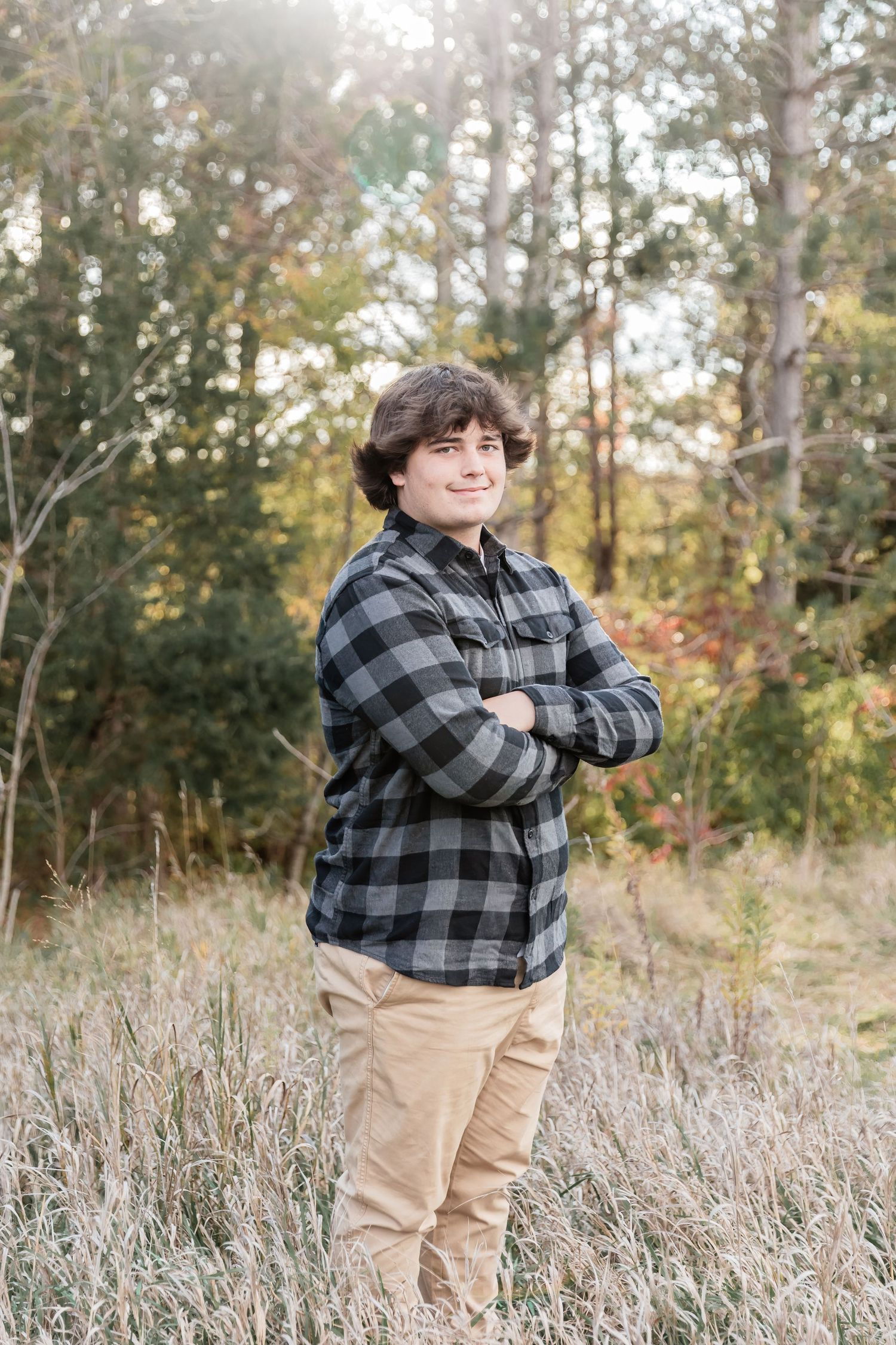 Young teenage boy stands with arms crossed in front of a pine tree forest
