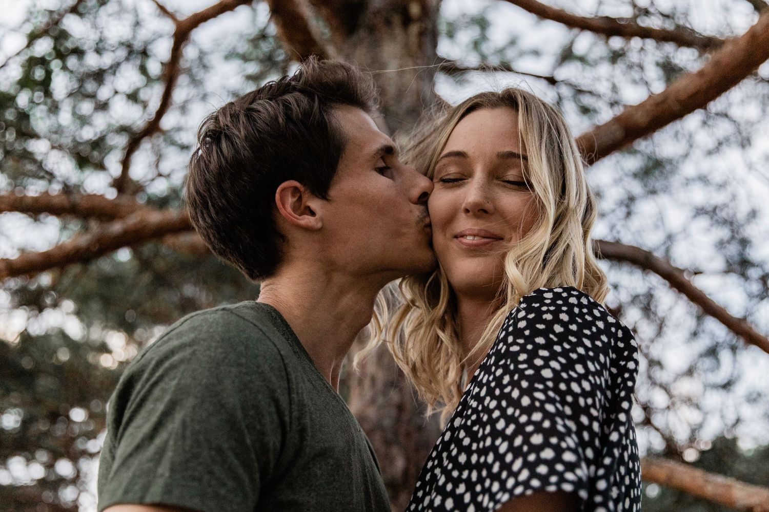 young woman enjoys a kiss on the cheek from her boyfriend under a pine tree
