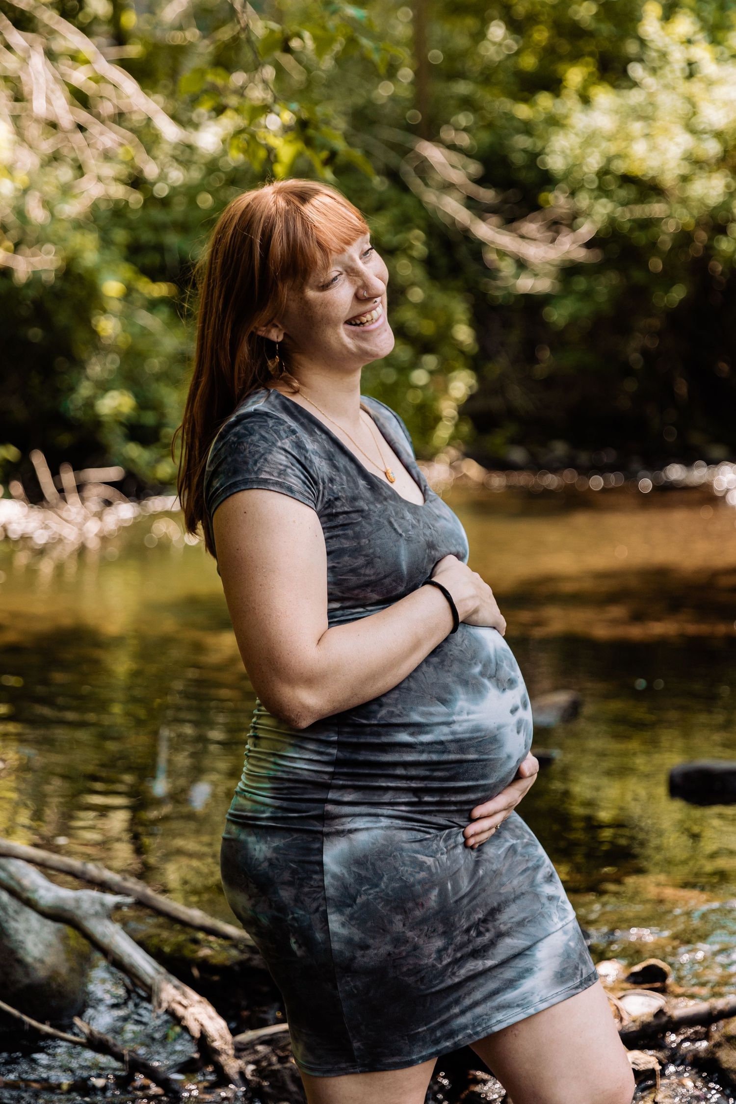 Pregnant red headed woman smiles and holds her baby bump while she stands in a babbling creek