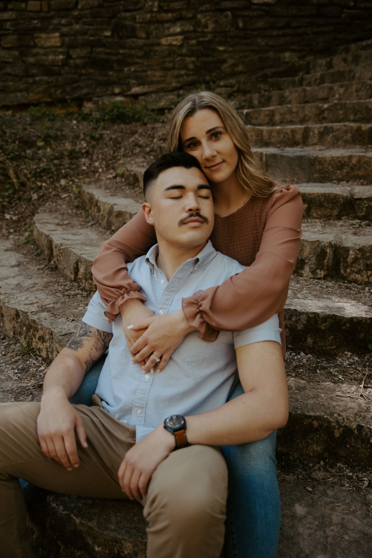 Young mane leans back into his fiance on a stony staircase as she wraps her arms around him