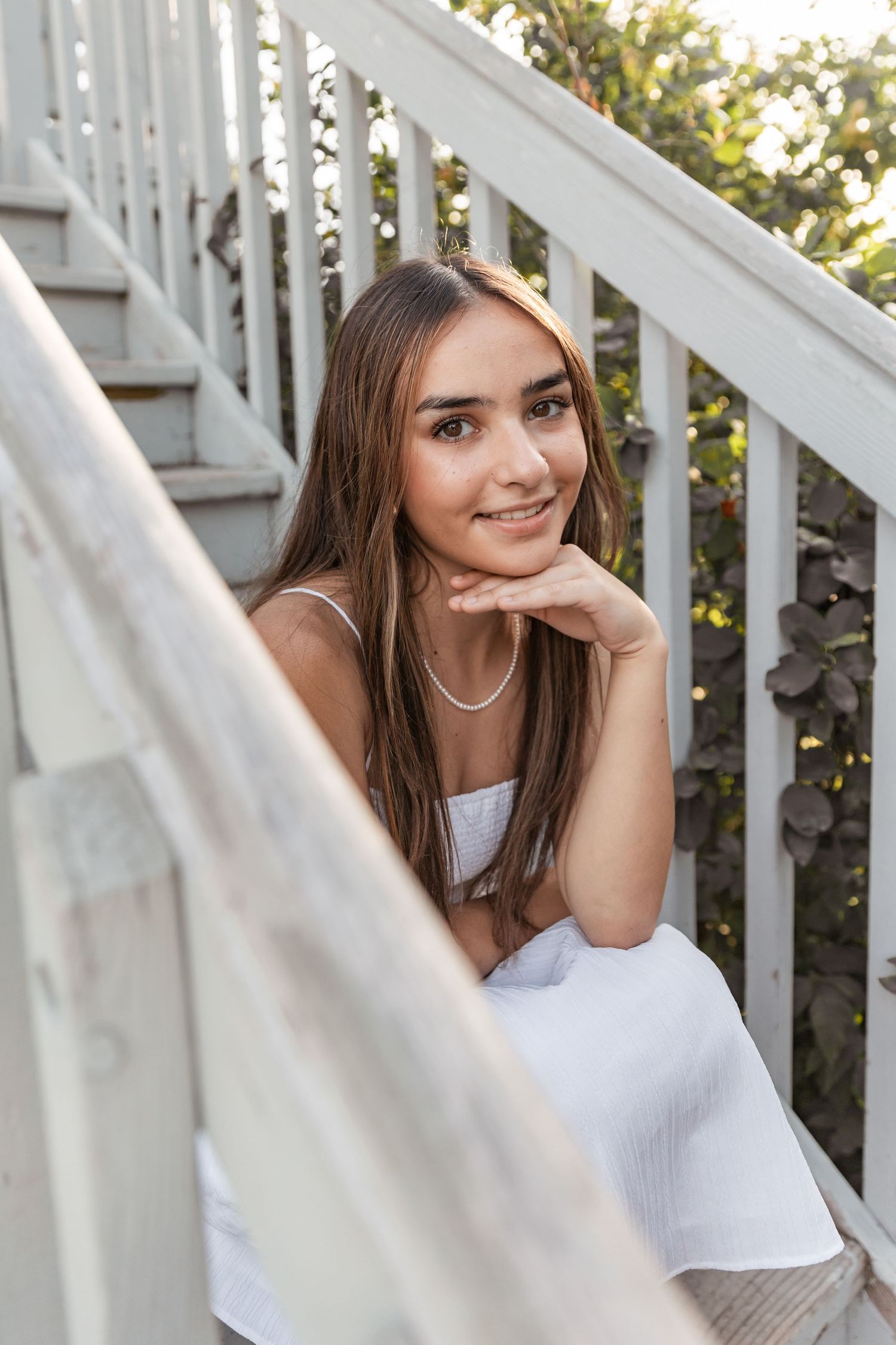 Maya sits on a white wooden staircase in a flower garden  with a golden glow of sunlight behind her