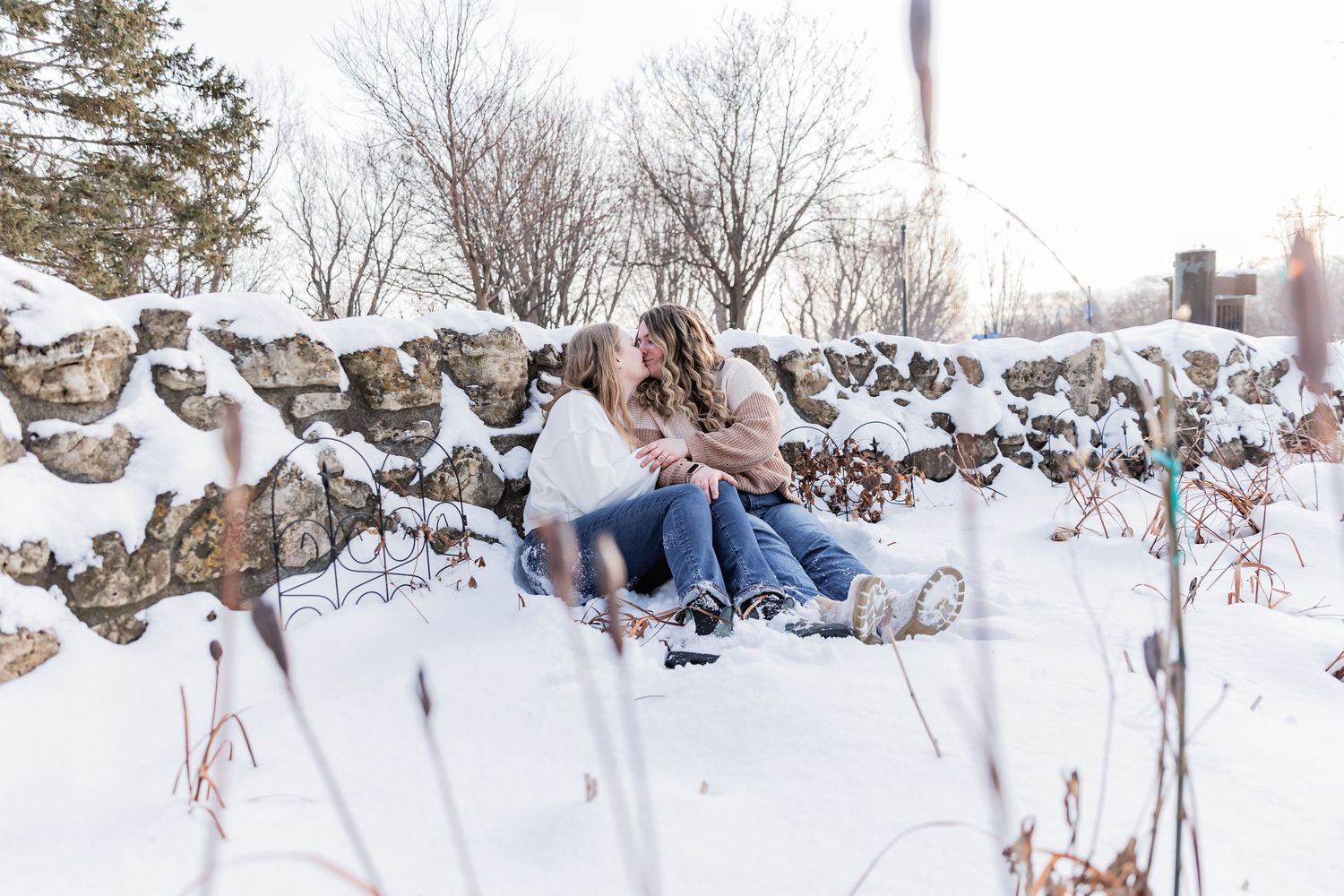 Young lesbian couple sits in the snow against a snow covered rock wall as they cuddle with each other