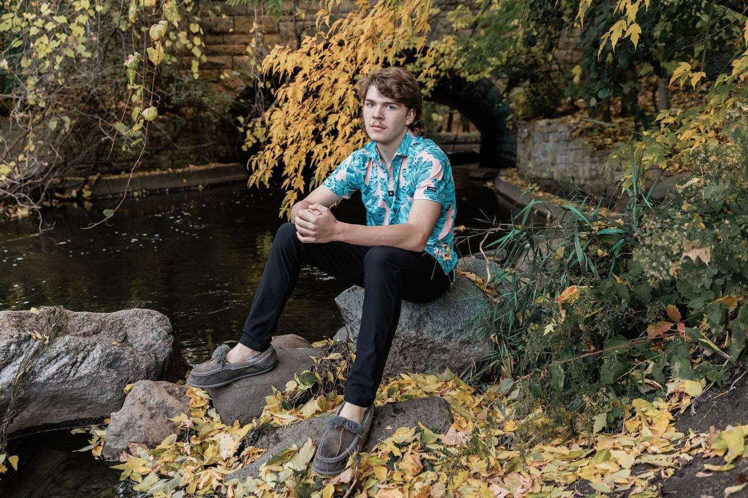 Minnehaha Academy basketball player sits on large rocks in the creek above Minnehaha Falls for his senior photos