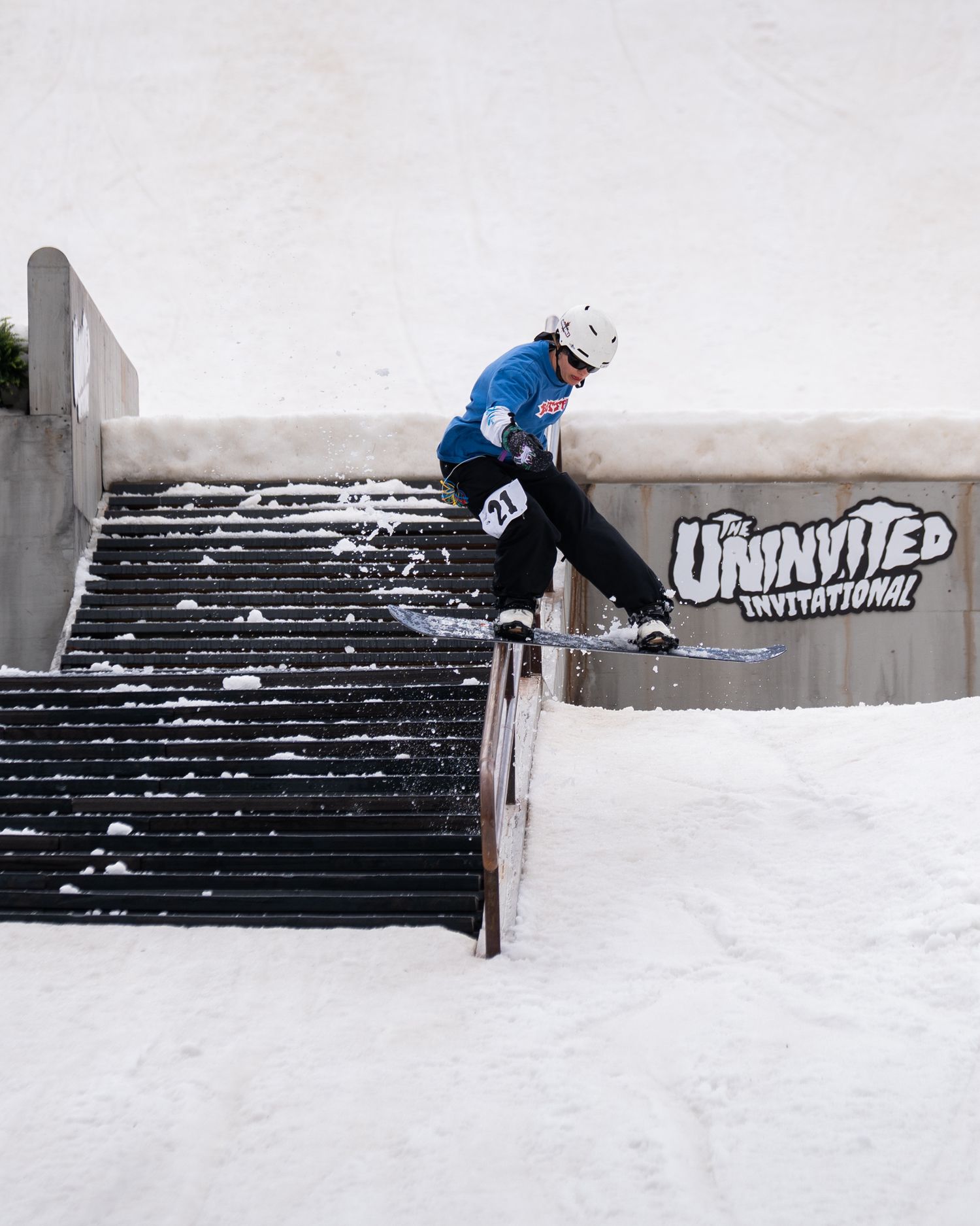 Snowboarder in blue jacket grinding a stair rail at The Uninvited Invitational competition.
