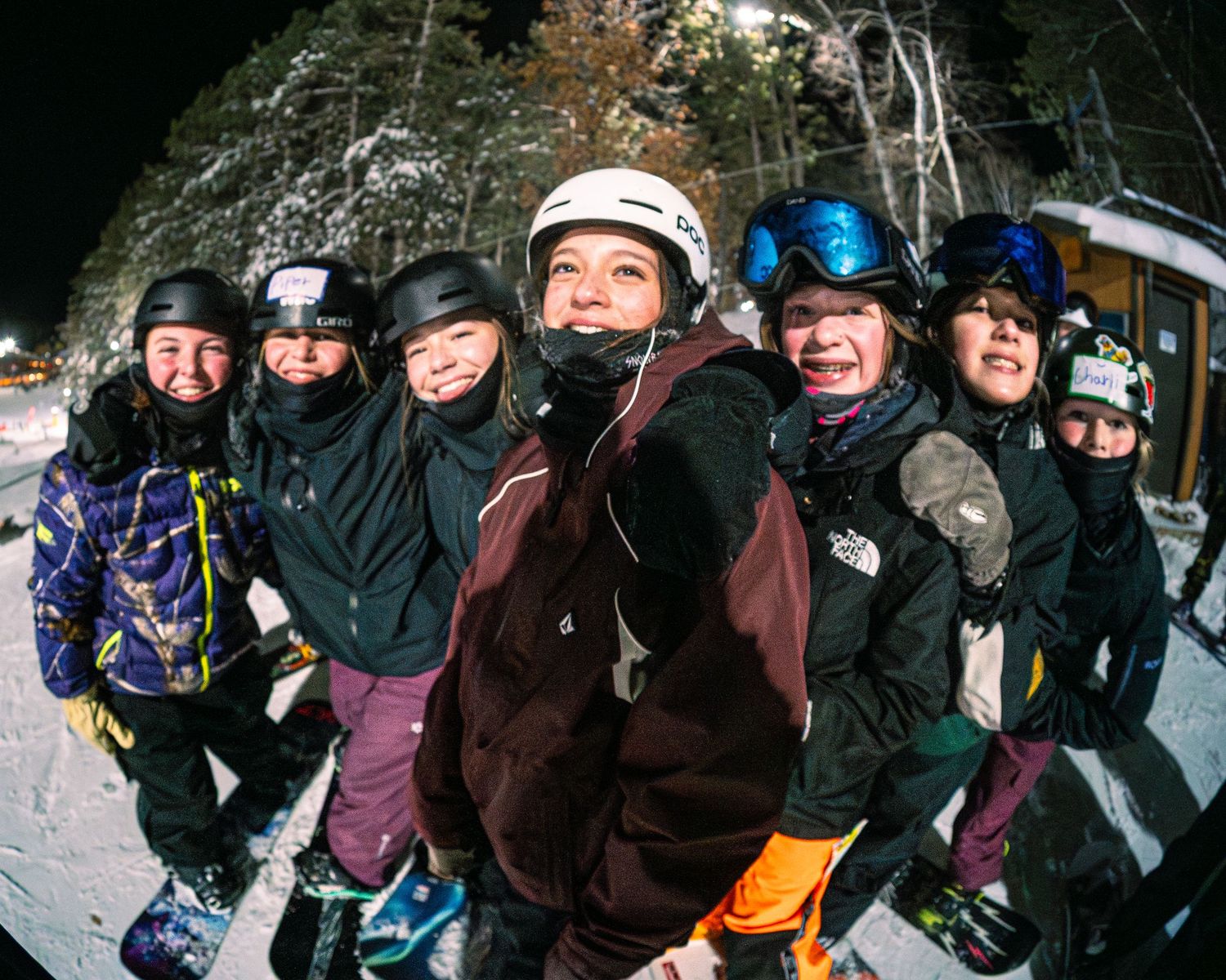 Group of smiling snowboarders posing together at a night ski resort, wearing helmets and winter gear.