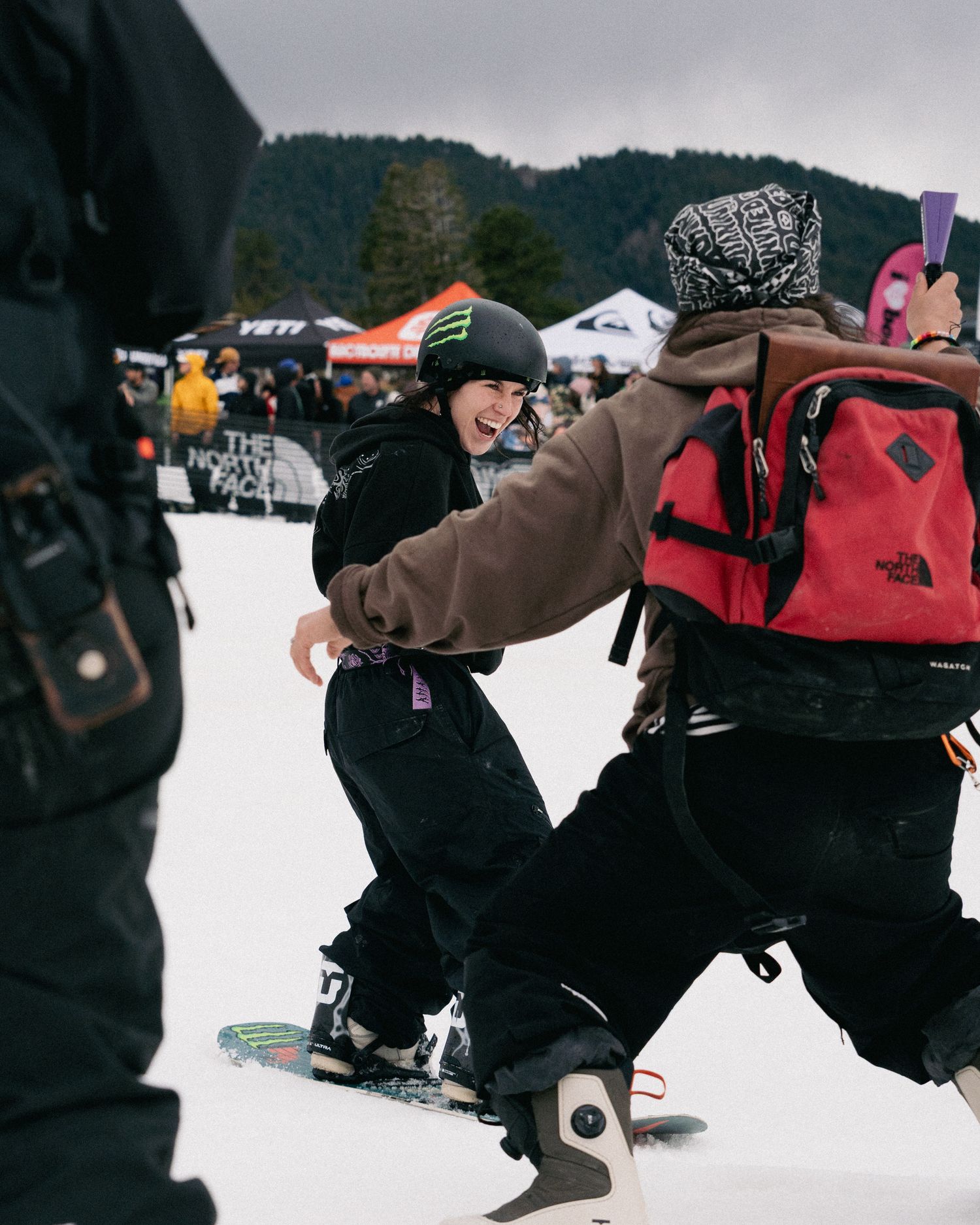 Snowboarder in Monster Energy helmet reaches out to greet someone at an outdoor winter snowboard competition.