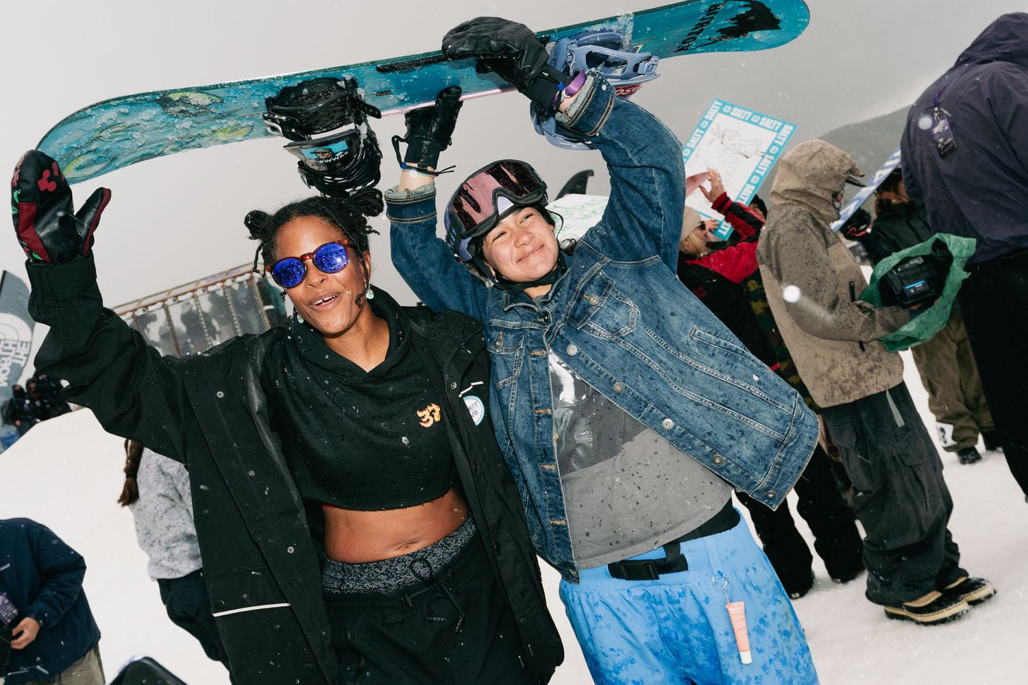 Two snowboarders celebrate holding board overhead at winter snowboard competition event.