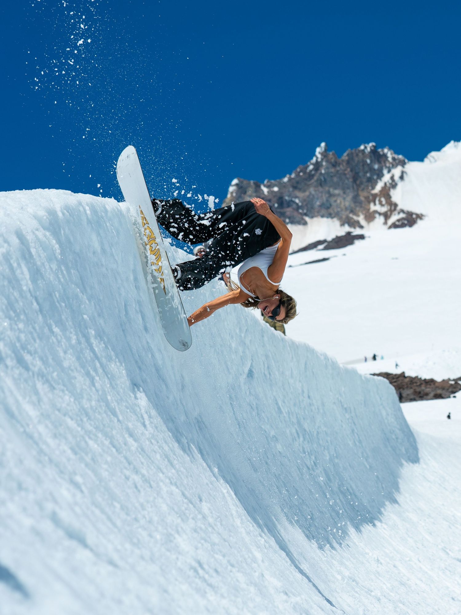 Snowboarder performing aerial trick off snow wall with mountain peak in background.