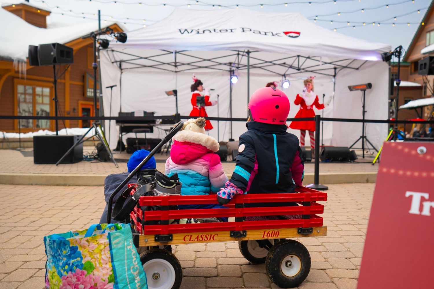 Holiday performers in festive costumes sit on a red wagon at a winter event.