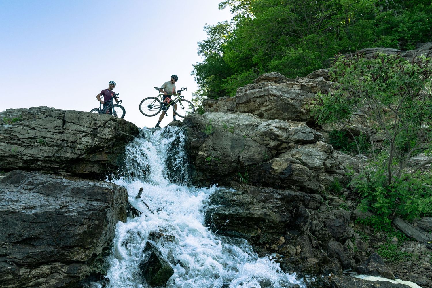 Two mountain bikers carry their bikes across rocky terrain above a rushing waterfall surrounded by lush green trees.