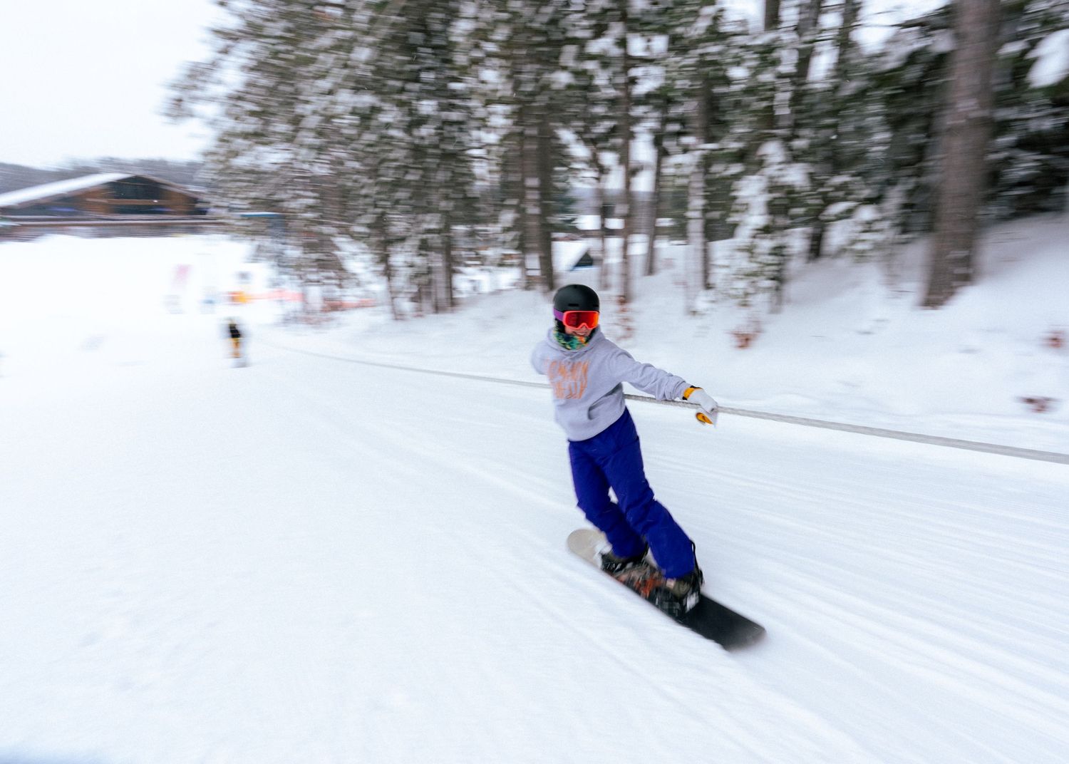 Snowboarder in red mask and blue pants carving down a snowy ski slope with trees in the background.