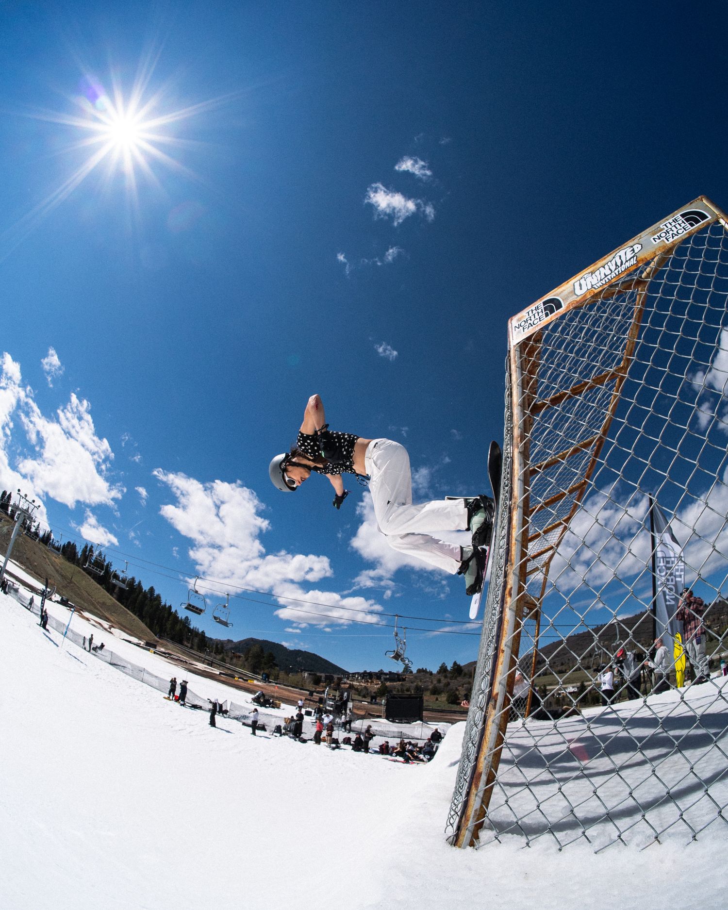A snowboarder performs a high aerial trick beside a net under a bright sun at a ski resort.