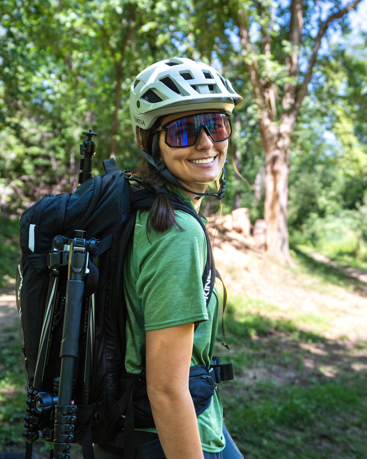 A mountain biker in a green shirt and helmet carries photography gear while riding trails in a wooded forest setting.