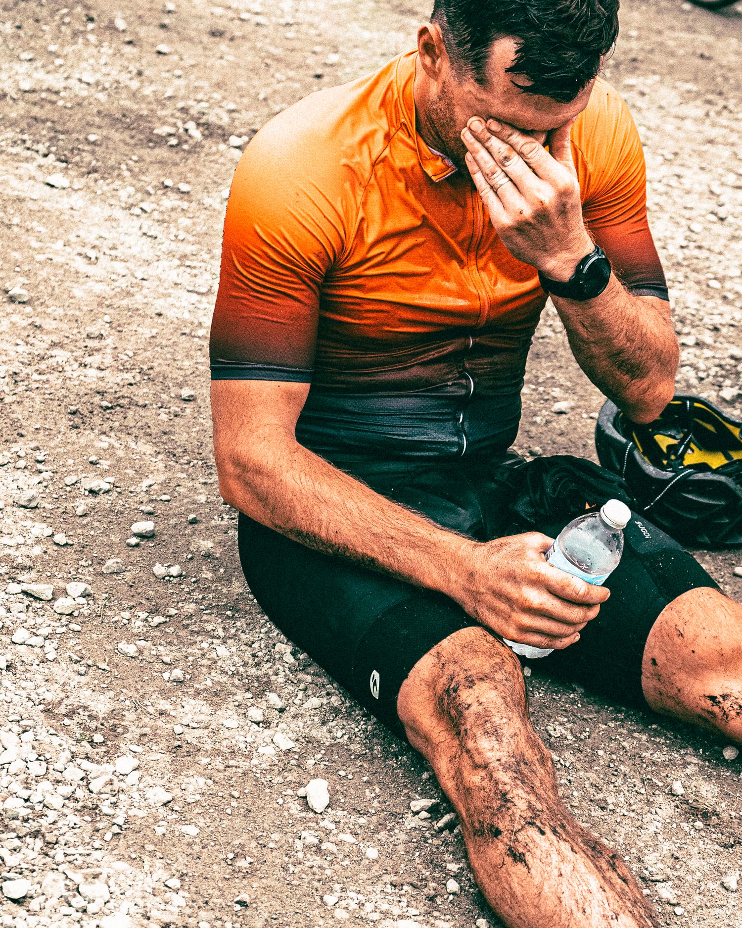 Exhausted cyclist in orange jersey sitting on gravel, holding water bottle after a muddy race.