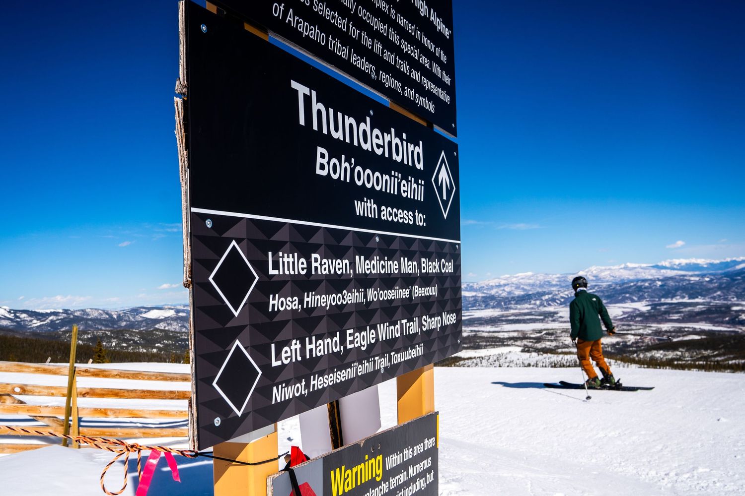 Ski run sign marking Thunderbird trail with skier passing by in winter mountain landscape.