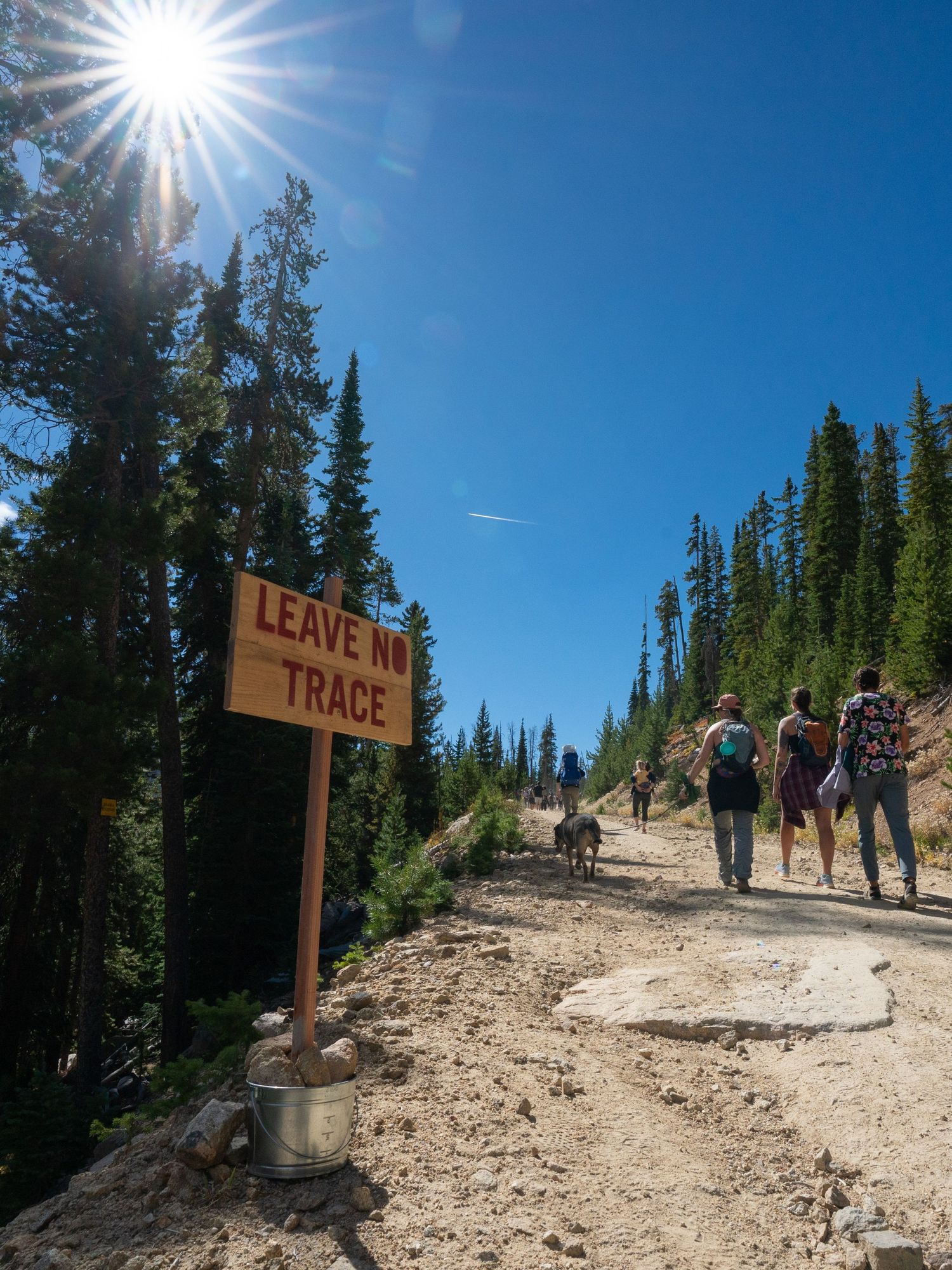 Sun-drenched mountain trail with Leave No Trace sign and hikers ascending path.