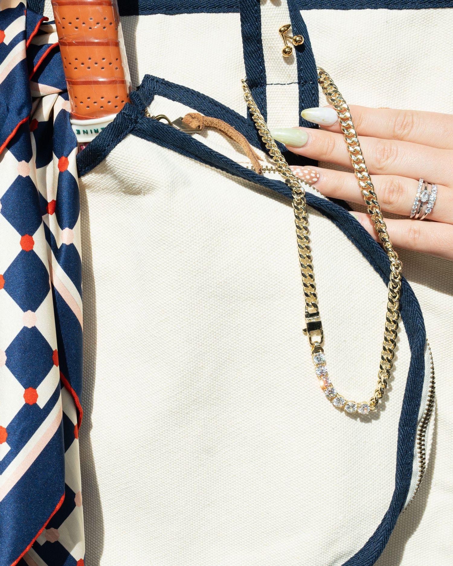 Woman's hand with manicured nails holding a gold chain necklace over a cream jacket with silk scarf.