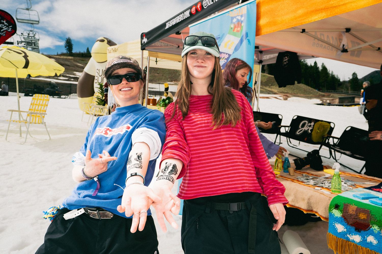 Two skiers pose and smile at a sunny outdoor ski resort event with colorful tents in the background.