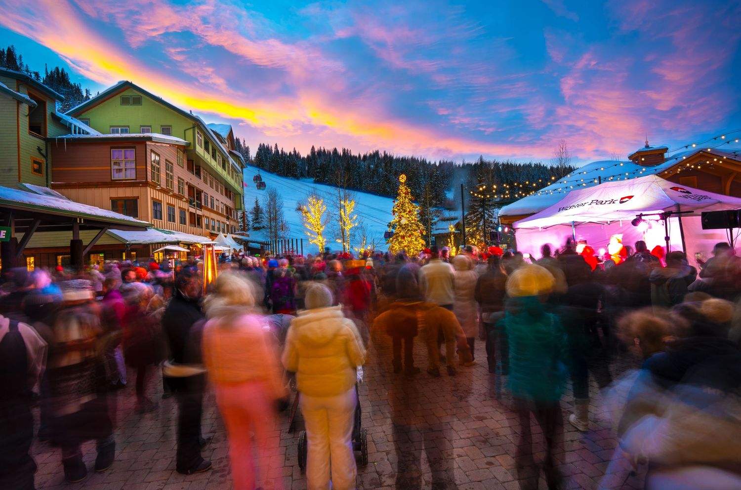 Crowd gathers at winter resort village plaza during dramatic sunset with lit buildings and trees.