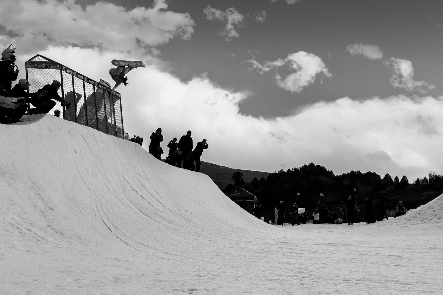Snowboarder performing a trick on a halfpipe ramp at a snow park, with spectators watching in black and white.