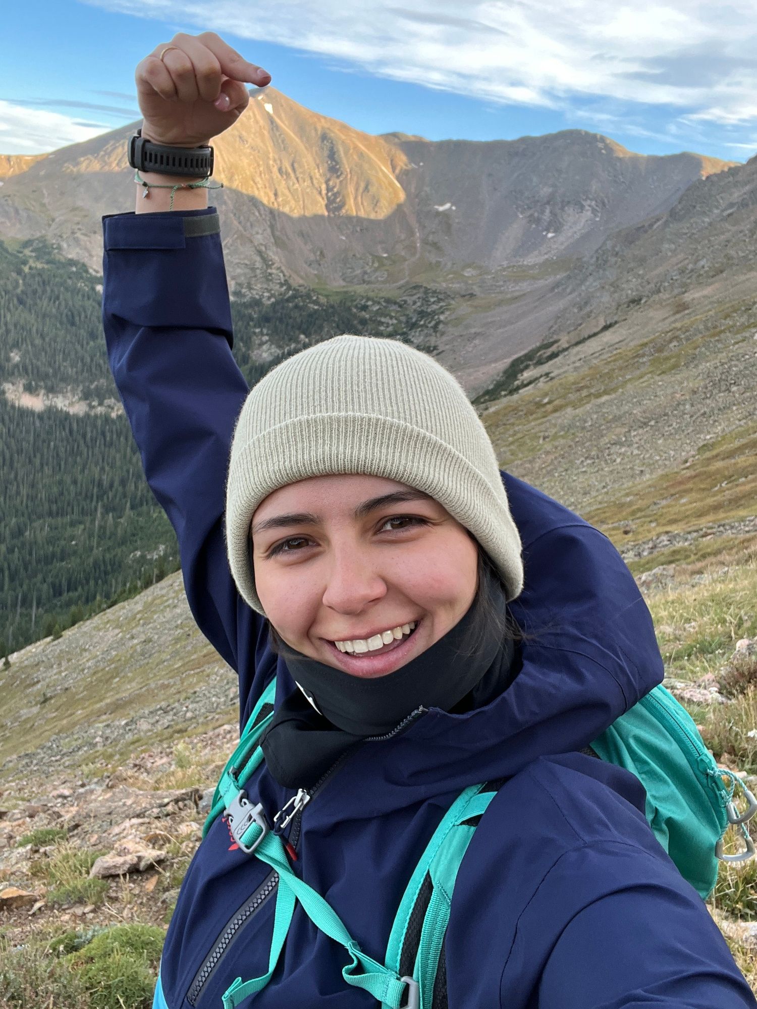 A hiker in winter clothing stands on a mountain trail with scenic rocky peaks and evergreen forests in the background.