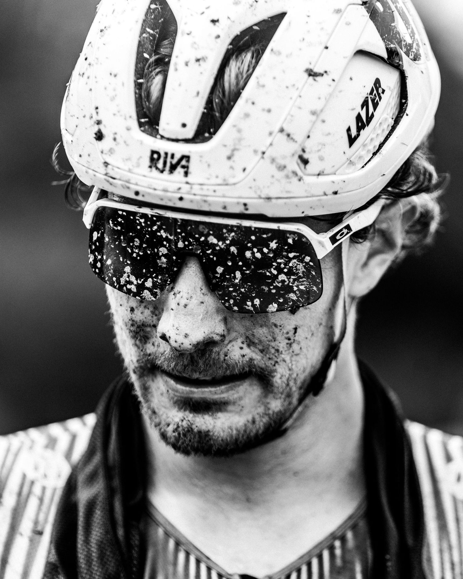 Black and white close-up of a muddy cyclist wearing a Lazer helmet and sunglasses after a race.