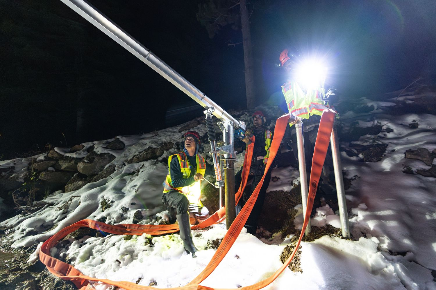 Night ski slope preparation with snow-making equipment and bright lights illuminating snowy terrain.