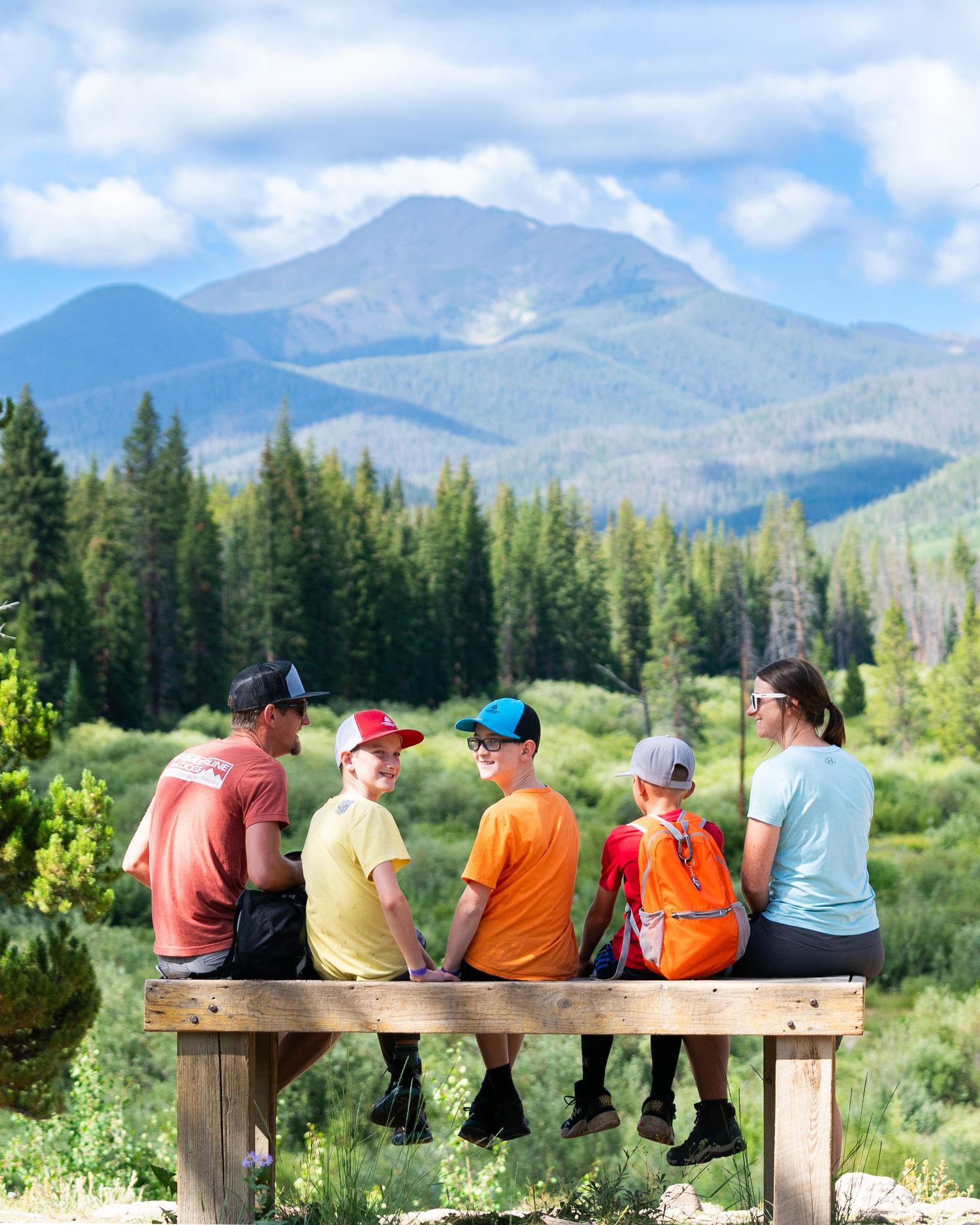 Group resting on wooden bench overlooking majestic mountain landscape with evergreen forests.