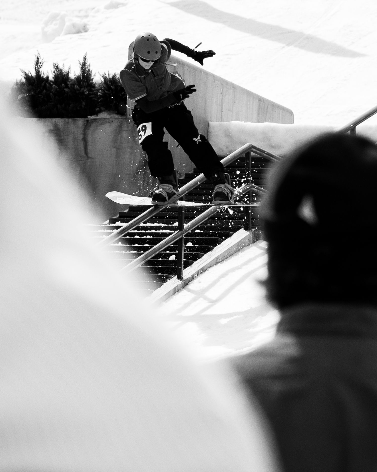 Snowboarder grinding a rail at a snowy terrain park in black and white action shot.