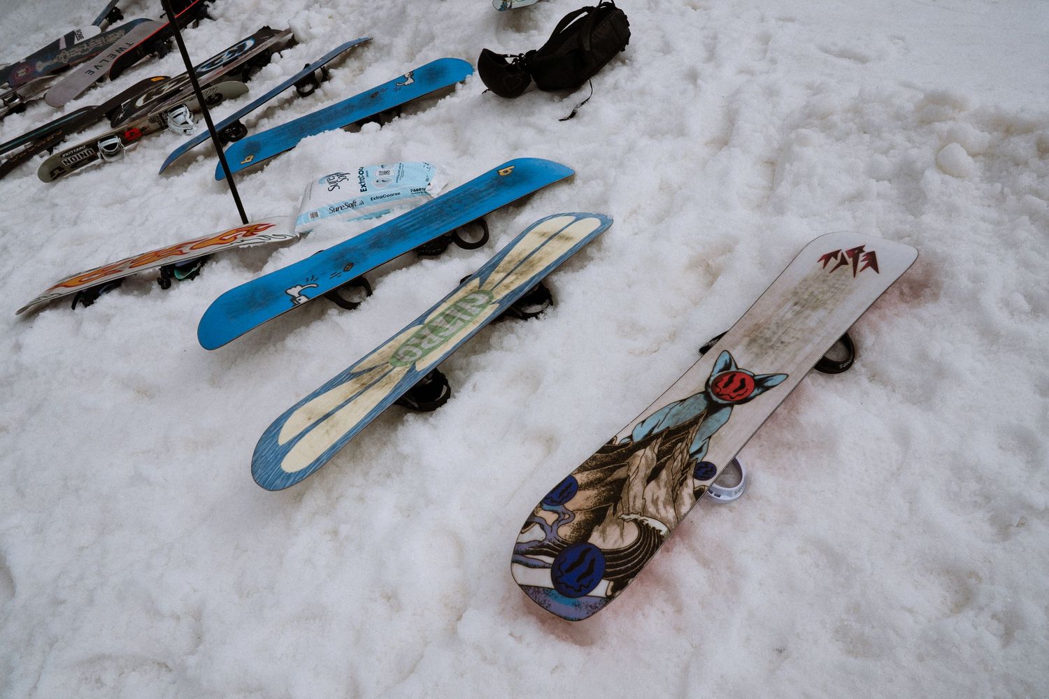 Three colorful snowboards laid flat on white snow at a ski resort, with ski poles and equipment visible in the background.